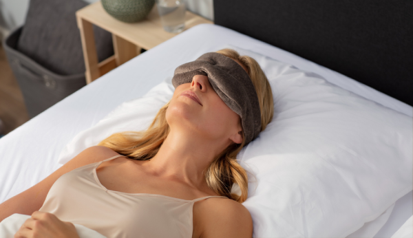 Woman wearing a sleep mask lying on a bed in a bedroom setting.