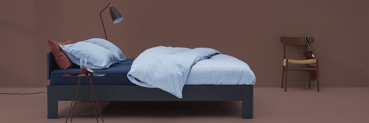 Bed with red mattress cover and gray pillows, wooden nightstand, and chair against a green wall.