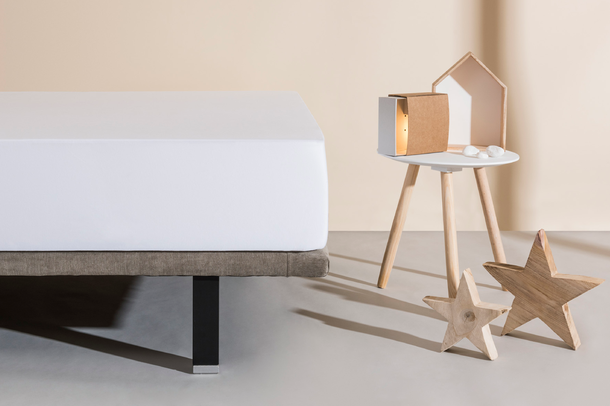 Bed with a white mattress in a minimalistic room with a beige wall and wooden nightstand.